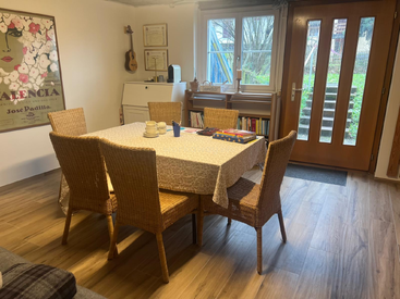 A cozy dining room with six wicker chairs, wooden floor, large window, wall art, bookshelf, and table set for coffee. Guitar and certificates decorate the wall.