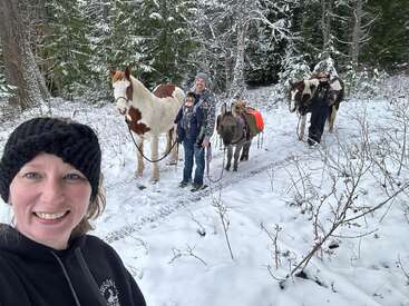 A smiling woman takes a selfie in a snowy forest. Behind her, two adults, two horses, a child, and a donkey enjoy a wintery walk together.
