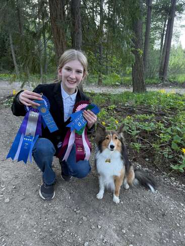 Ein lächelndes Mädchen kniet auf einem Waldweg und hält stolz mehrere Preisbänder in den Händen. Neben ihr sitzt ein flauschiger Hund, und beide genießen ihren Erfolg im Freien.