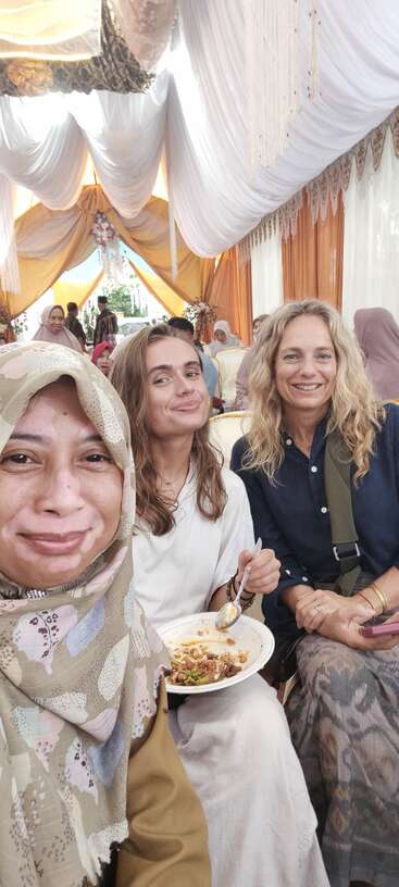 Three women smile for a selfie at a festive event with elaborate drapes, surrounded by others. One enjoys food, capturing a joyful multicultural gathering moment.