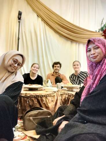 Five people sit together on the floor around a table with food, smiling. The background features draped golden fabric, creating a warm and festive atmosphere.