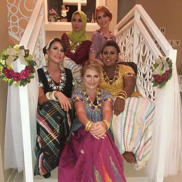 The image depicts five women posing on a white staircase, adorned with flowers and tulle, showcasing their diverse traditional attire. The women are seated in a harmonious arrangement.