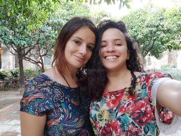 The image shows two women taking a selfie in an outdoor setting, with trees and buildings visible in the background. They are both wearing floral tops.