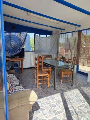 The image depicts a cozy room with a dining table, TV, and couch, featuring blue trim and a stone floor, bathed in natural light from the large windows.