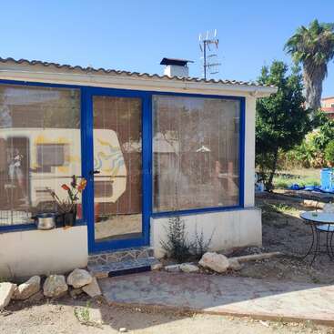 The image depicts a small, white building with blue-framed windows and a door, situated in a yard with a table and chairs, surrounded by trees and a clear blue sky.