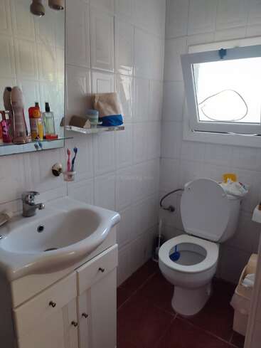 The image depicts a compact bathroom with white tiles, featuring a sink, toilet, and window, with a mirror above the sink and a small shelf on the wall.