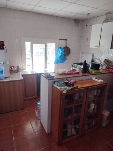 The image depicts a kitchen with white tiled walls and floor, featuring wooden cabinets, a stove, and various kitchen items, with a window and door leading to the outside.