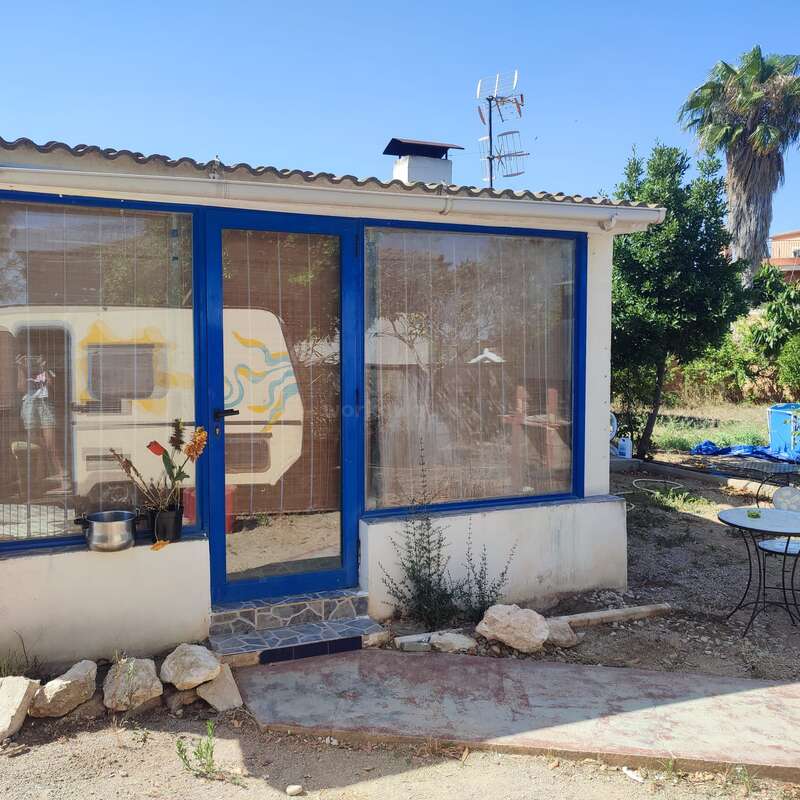 The image depicts a small, white building with blue-framed windows and a door, situated in a yard with a table and chairs, surrounded by trees and a clear blue sky.