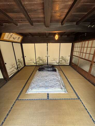 Esta imagen muestra una habitación tradicional japonesa con tatamis, puertas correderas de shoji, un futón en el centro y una decoración minimalista con techo de madera.