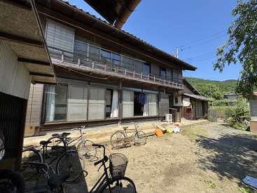 Se trata de una casa tradicional japonesa de dos plantas con puertas correderas y un balcón; varias bicicletas aparcadas en el exterior, un cielo azul claro y la vegetación circundante crean un ambiente tranquilo.
