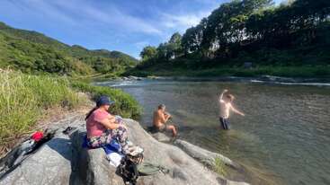 Tres personas disfrutan de un día soleado junto al río; una está sentada en una roca, otra descansa a la orilla del agua y la tercera se adentra en el agua. Todo ello rodeado de una exuberante naturaleza.