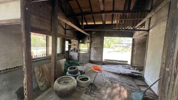This image shows an old, abandoned, and partially damaged room with wooden beams, large pots, a green rake, an orange scoop, and sunlight streaming in.