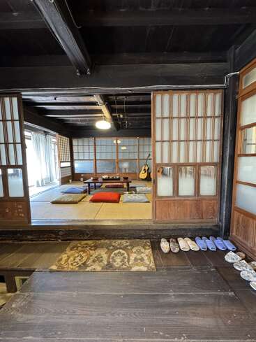 A traditional Japanese room with tatami flooring, sliding shoji doors, low table, colorful floor cushions, slippers neatly lined up, guitar in the corner, cozy ambiance.