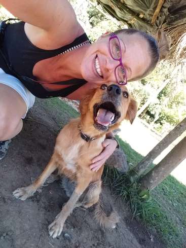 The image shows a woman kneeling beside a brown dog with its mouth open, posing for a selfie in a park-like setting with grass and trees.