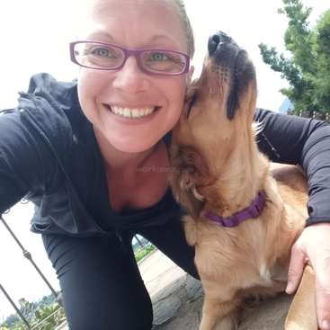 The image shows a woman in black clothing posing with a golden retriever, with the dog\'s nose touching her cheek, set against a bright outdoor background.