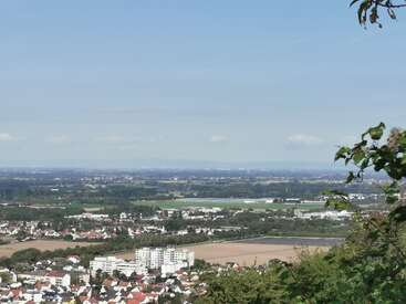 The image depicts a small town with white buildings and red roofs, surrounded by trees and fields, set against a blue sky with a few clouds.