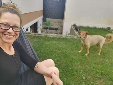 Eine lächelnde Frau mit Brille sitzt im Freien auf einer Schaukel, während ein hellbrauner Hund in der Nähe auf dem Gras steht. Im Hintergrund sind ein Gebäude und ein Garten zu sehen.