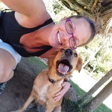 Das Bild zeigt eine Frau, die neben einem braunen Hund mit offenem Maul kniet und für ein Selfie in einer parkähnlichen Umgebung mit Gras und Bäumen posiert.