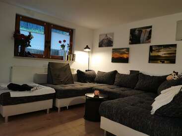 Ein gemütliches Wohnzimmer mit einem großen Sektionssofa, Wandkunst, einer Stehlampe, einem kleinen runden Tisch, Blumenvasen am Fenster und einem Holzfußboden.