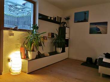 Ein gemütliches Wohnzimmer mit Holzböden, Topfpflanzen, modernen Regalen, Büchern, einer leuchtenden Lampe, Wandkunst, einem Fenster und ordentlicher Dekoration, die ein warmes Ambiente schafft.