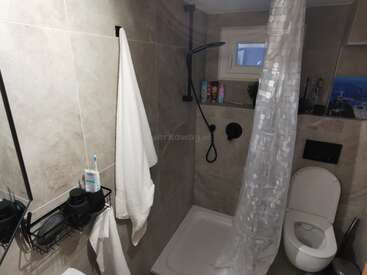 Dieses Bild zeigt ein modernes Badezimmer mit beigen Fliesen, einer wandmontierten Dusche, einem weißen Vorhang, Handtuch, Toilette, Toilettenartikeln auf Regalen und einem kleinen Fenster darüber.