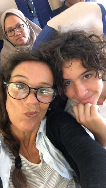 A woman and a boy take a close-up selfie, smiling, while a woman in a headscarf sits behind them, looking serious on a plane.