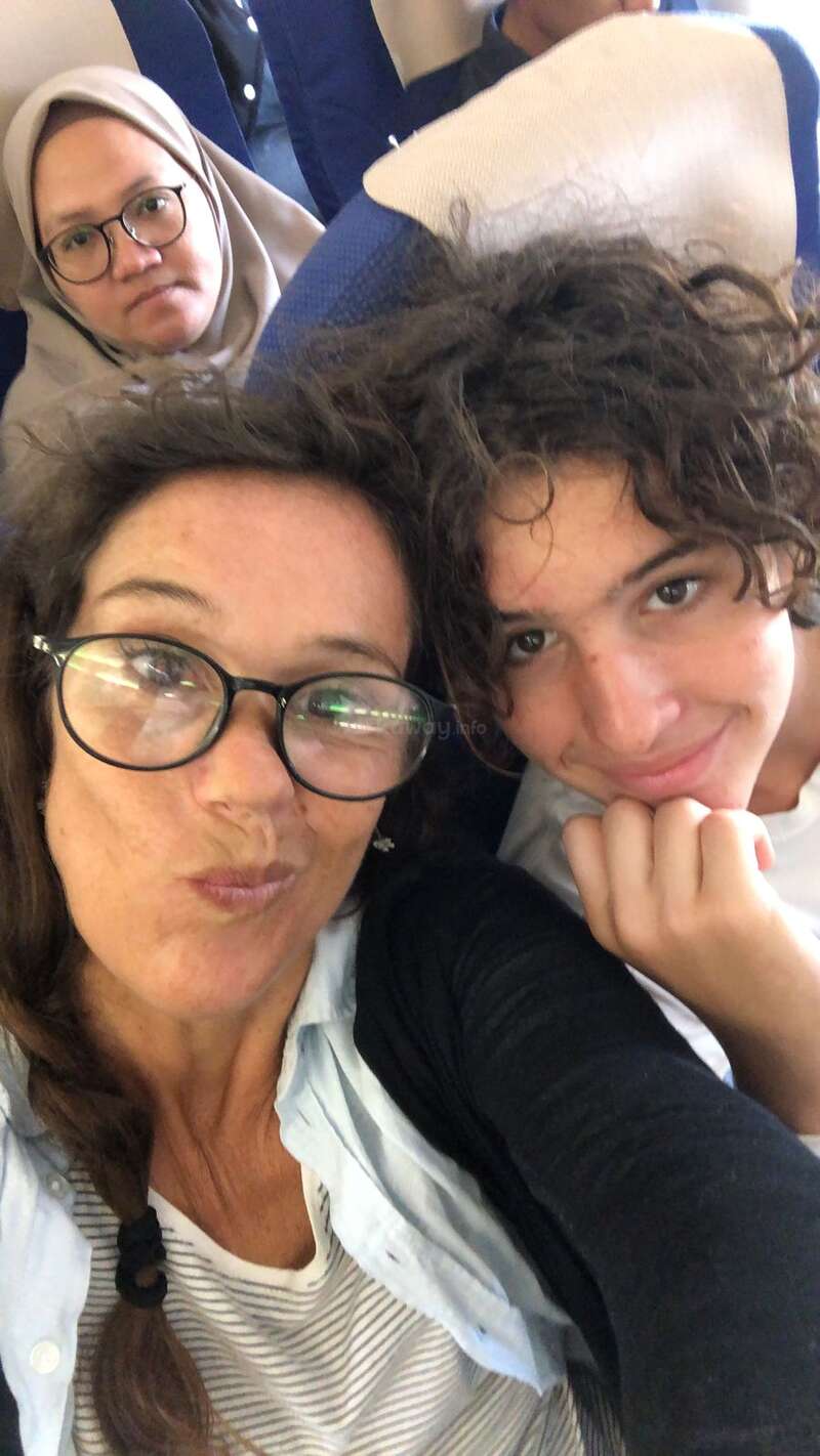 A woman and a boy take a close-up selfie, smiling, while a woman in a headscarf sits behind them, looking serious on a plane.