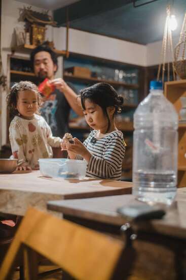 Two young children stand at a wooden table, focused on food preparation. An adult observes them from behind. The kitchen appears cozy and warmly lit.