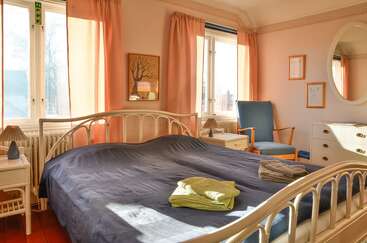 Este quarto aconchegante tem uma cama grande com uma colcha azul-escura, cortinas em tons pastéis, luz do sol entrando pela janela, uma cadeira azul e toalhas bem dobradas.