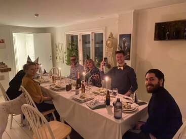 Seis pessoas usando chapéus de festa estão sentadas ao redor de uma mesa de jantar, saboreando comida e bebidas. Velas, decorações festivas e um ambiente acolhedor e amigável tomam conta da sala.
