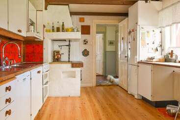 Esta cozinha aconchegante tem piso de madeira, armários brancos, azulejos vermelhos no revestimento da parede, decoração rústica, muita luz natural e um cachorrinho descansando perto da entrada do corredor ao fundo.