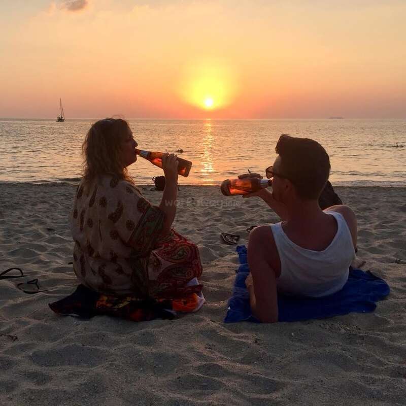 Um casal está sentado na praia ao pôr do sol, tomando cerveja juntos. O sol se põe sobre o mar calmo, lançando um brilho quente e tranquilo.
