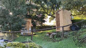Esta imagen muestra una zona exterior cubierta de césped con dos pequeñas construcciones de madera entre los árboles, la luz del sol filtrándose a través de las hojas y una barandilla rústica de madera que sigue el terreno inclinado.