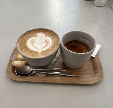 Auf einem Holztablett stehen ein Cappuccino mit Latte Art, ein Espresso, zwei kleine Kekse und zwei Löffel, die alle ordentlich auf einem grauen Tisch liegen.