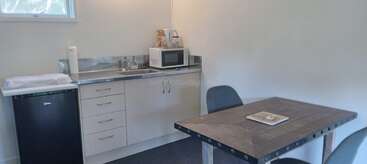 Esta imagen muestra una pequeña cocina con un mini frigorífico, fregadero, cajones, microondas, hervidor de agua y una mesa de comedor cuadrada de madera con dos sillas cerca de una ventana.