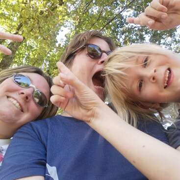 La imagen muestra a tres personas posando para un selfie, dos de ellas haciendo gestos con las manos y una con gafas de sol, sobre un fondo de árboles.
