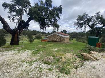 A small rustic stone building with a red roof sits amid green grassy land and large trees under a cloudy sky, surrounded by natural countryside scenery.