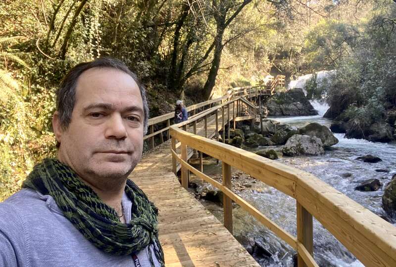 A man takes a selfie on a wooden walkway beside a flowing stream, surrounded by lush green forest. Another person walks ahead, enjoying nature.