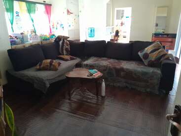 A cozy living room with a dark L-shaped sofa, colorful pillows, a small wooden coffee table, bright curtains, toys, and an open view to the kitchen.