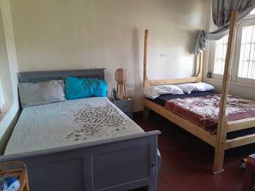 This image shows a simple bedroom with two beds, one gray and one wooden. There is a nightstand between them with a lamp and window nearby.