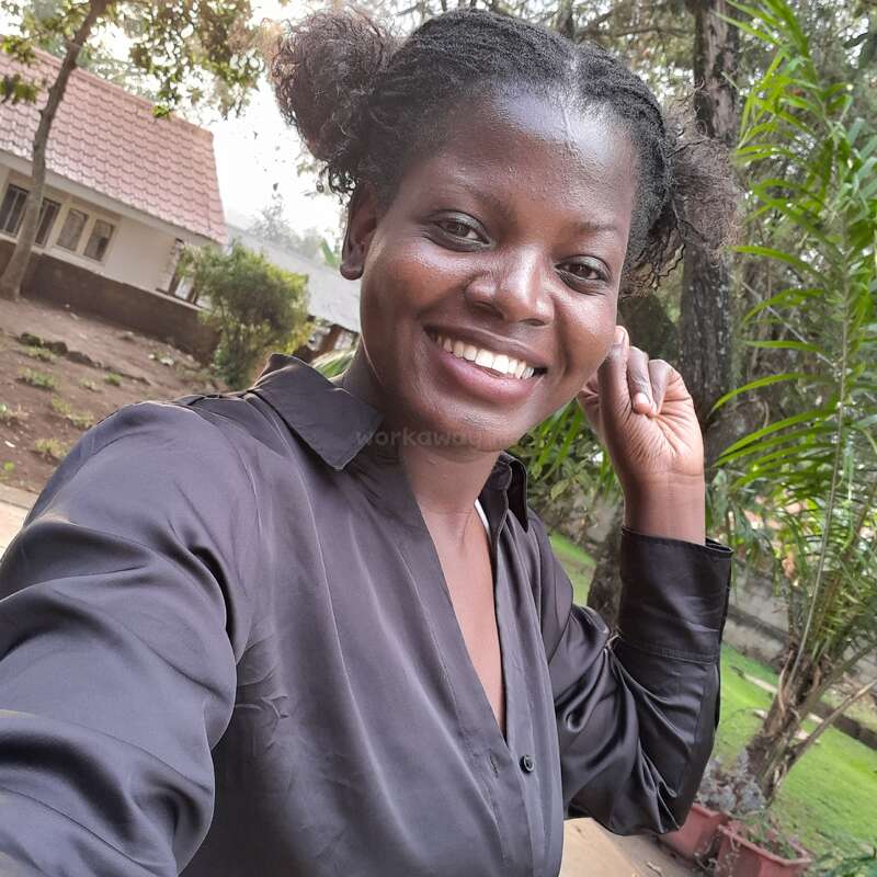 A smiling woman takes a selfie outdoors, wearing a black shirt. Behind her are green plants, trees, and a house with a tiled roof. Relaxed atmosphere.