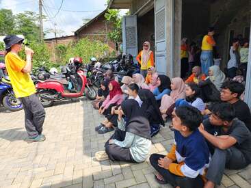 A group of young people sit attentively outdoors, listening to a speaker in a yellow shirt. Motorbikes are parked behind them, and others stand nearby.