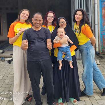 A cheerful group of six people, including a smiling baby, pose together outside a building. Four women wear yellow shirts, one man gives thumbs up. Dated July 2025.
