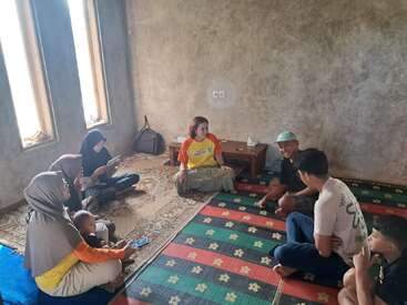 A group of people sits on colorful mats in a simple room with bare walls. Some are chatting, others using phones. One woman holds a baby.