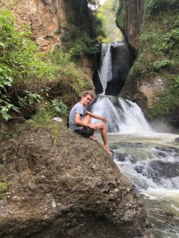 A smiling person sits barefoot on a rock beside a rushing waterfall surrounded by lush greenery, rocky cliffs, and cascading water in a serene natural setting.