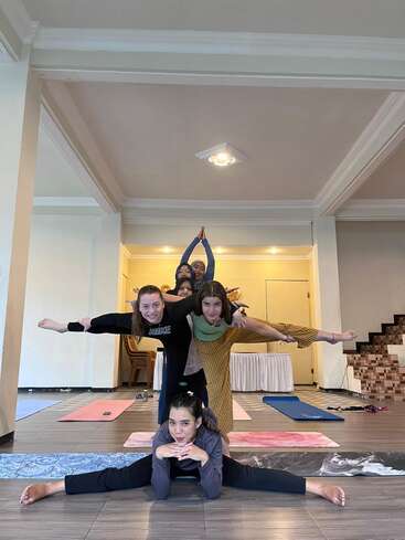 Five women are stacked in a creative yoga pose inside a spacious room, smiling and balancing on each other. Yoga mats and relaxed atmosphere surround them.
