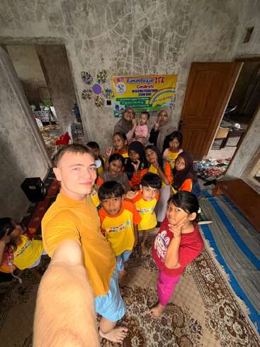 A group of children and adults pose smiling for a selfie inside a room with decorated walls, a carpet, and a colorful educational poster in the background.