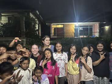 A group of smiling children and two adults pose together outdoors at night in front of a brightly lit house, making peace signs and enjoying the moment.