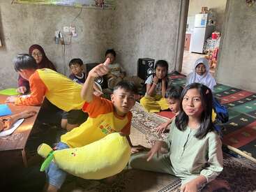A group of children sits together in a cozy room on carpets. Some are smiling, one gives a thumbs-up, and others appear relaxed and playful.