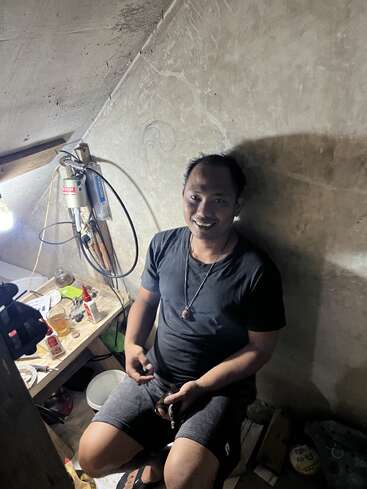 A man sits in a small, dimly lit room, smiling at the camera. Behind him is a cluttered table with tools, bottles, and various objects.
